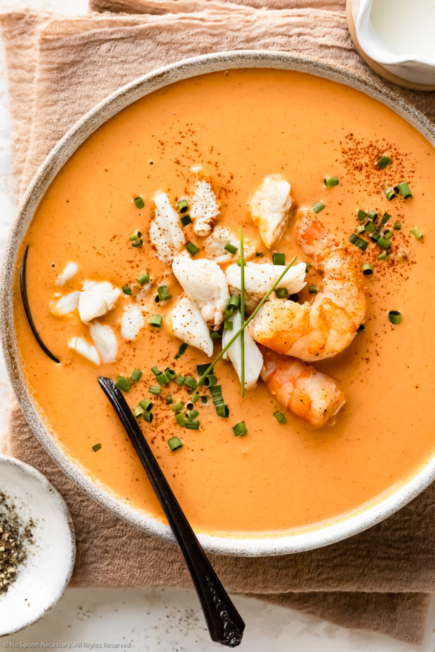 Overhead photo of Seafood Bisque garnished with lump crab and sautéed shrimp in a white soup bowl with a spoon inserted into the bisque and ramekins of fresh cracked pepper and cream next to the bowl.
