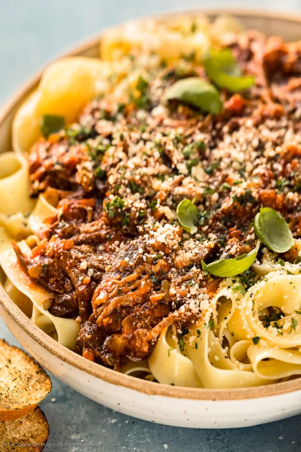 Angled, close-up photo of Slow Cooker Ragu Bolognese over papparelle pasta in a neutral colored serving bowl with slices of toasted baguette in front of the bowl and a ramekin of grated parmesan cheese blurred behind the bowl.