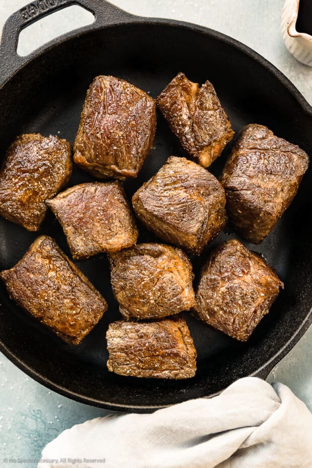 Overhead photo of seared, browned boneless beef short rib pieces in a cast iron pan with a ramekin of red wine next to the pan - photo of step 2 of the recipe.