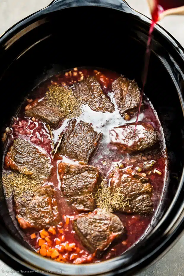 Slightly angled, overhead photo of red wine being poured over browned short ribs, vegetables, seasonings and crushed tomatoes in the pot of a slow cooker - photo of the second part of step 3 of the recipe.