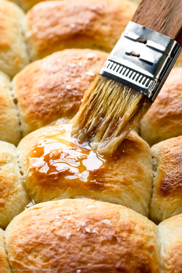 Angled, close-up photo of freshly baked soft yeast rolls being brushed with melted butter - step 8 of the recipe.