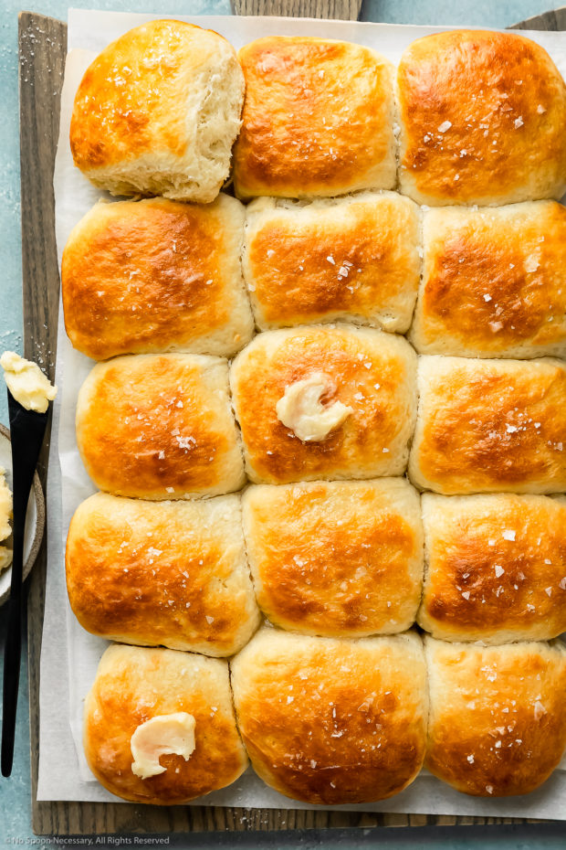 Overhead photo of freshly baked quick homemade dinner rolls slathered with butter on a large gray wood board with a ramekin of honey butter next to the rolls.