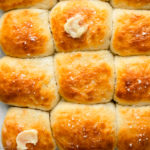 Overhead, landscape photo of freshly baked quick soft dinner rolls slathered with butter on a large gray wood board with a ramekin of honey butter next to the rolls.