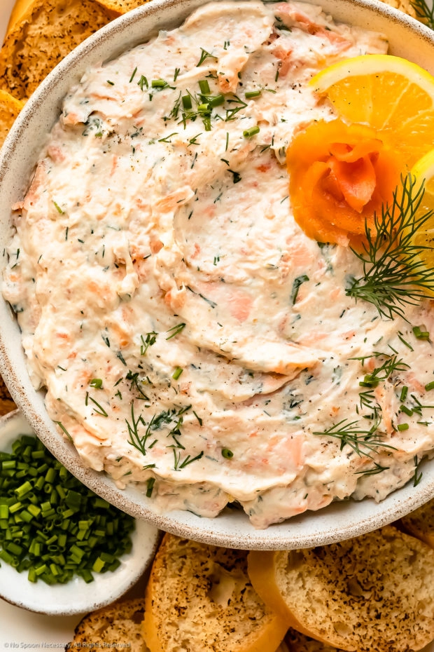 Overhead photo of smoked salmon spread topped with fresh dill in a white serving bowl with a ramekin of snipped chives and crusty slices of bread next to the bowl.