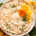Overhead photo of smoked salmon dip garnished with lemon slices and fresh dill in a white serving bowl with toasted slices of baguette surrounding the bowl.