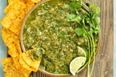Overhead photo of Salsa Verde garnished with lime wedges and fresh cilantro in a white serving bowl with a chip inserted into the green salsa.