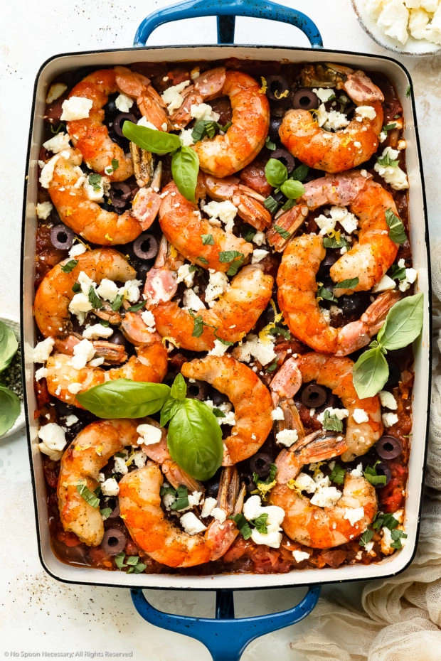 Overhead photo of baked Greek shrimp with tomatoes, olives and feta in a blue baking dish with ramekins of crumbled feta and fresh basil next to the pan.