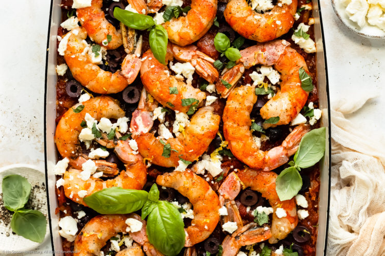 Overhead, landscape photo of baked Greek shrimp with tomatoes, olives and feta in a blue baking dish with ramekins of crumbled feta and fresh basil next to the pan.