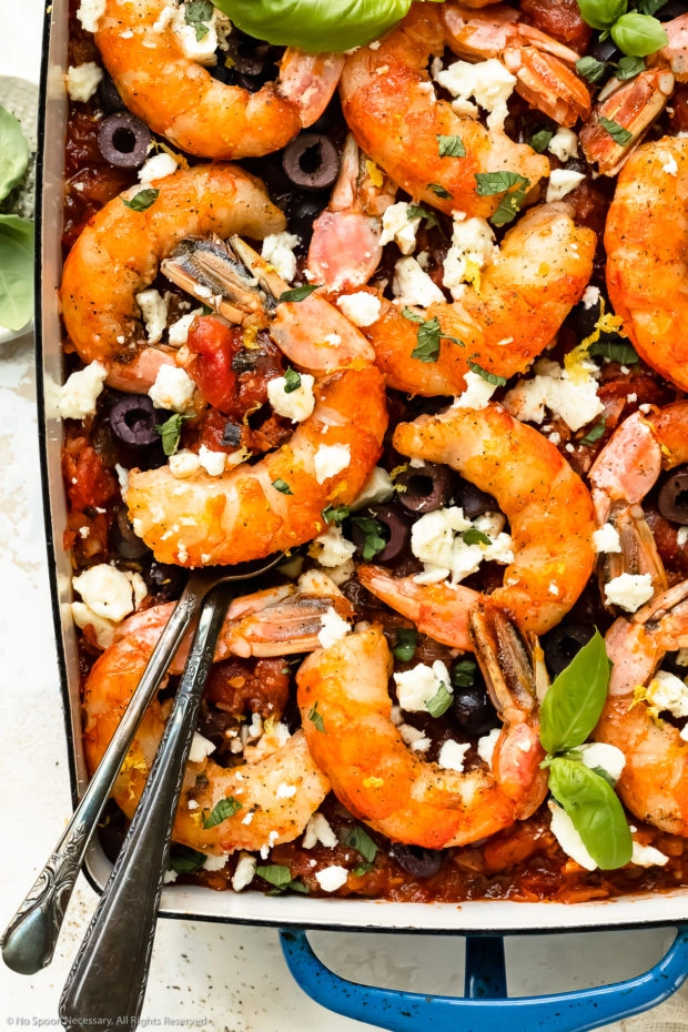 Overhead, close-up photo of Greek shrimp saganaki in a large blue baking dish with two serving spoons lifting up an individual shrimp with tomatoes.