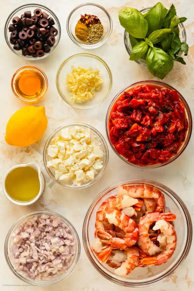 Overhead photo of shrimp, feta, garlic, lemon, olives, basil, red onion, and seasonings neatly organized on a kitchen counter.