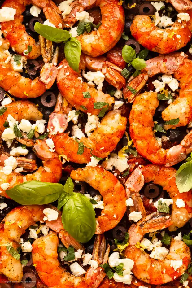 Close-up photo of cooked shrimp with crumbled feta cheese and fresh basil over a greek tomato mixture.