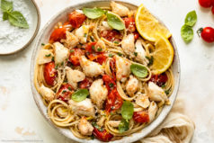 Overhead photo of a white bowl containing crab linguine in wine sauce garnished with fresh basil and lemon wedges.