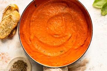 Overhead photo of Roasted Red Pepper Pasta Sauce in a large white saucepan with fresh basil leaves and cracked black pepper next to the pan.