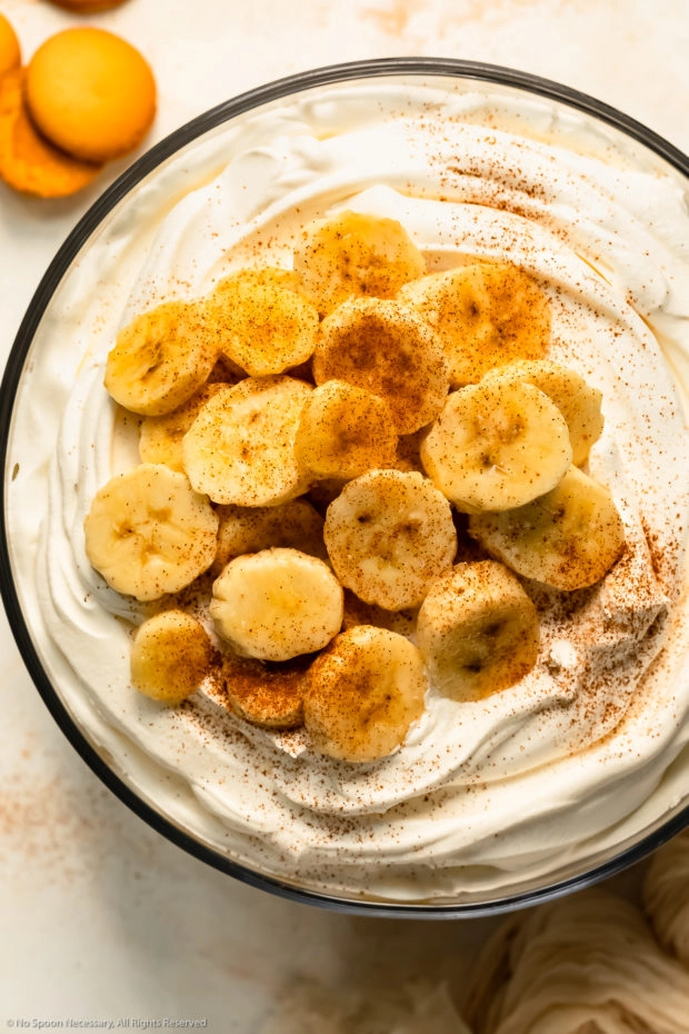 Overhead photo of fresh slices of bananas sprinkled with ground cinnamon on top of Cool Whip - photo of the top of a banana trifle.