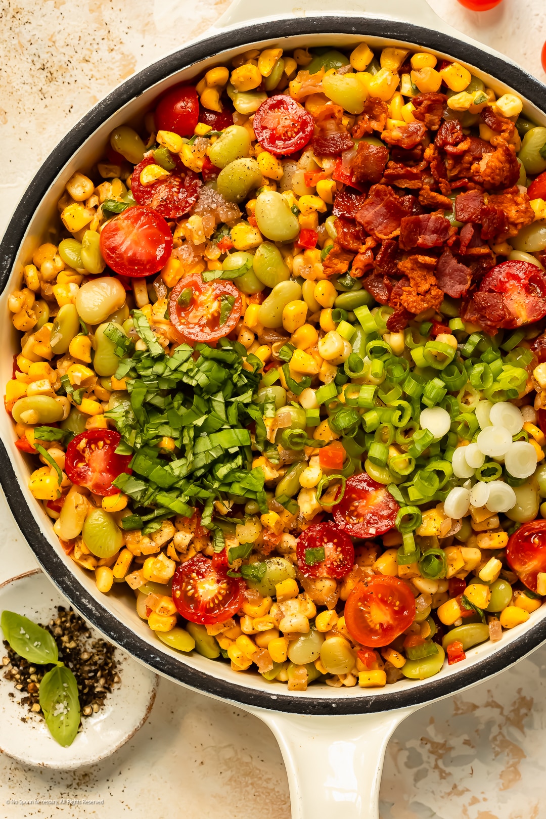 Overhead photo of a sweet corn, lima beans, tomatoes, crispy bacon, and fresh herbs in a white skillet.