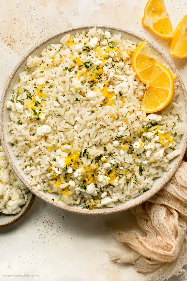 Overhead photo of Greek lemon rice garnished with crumbled feta, fresh herbs and lemon wedges in a white serving bowl.