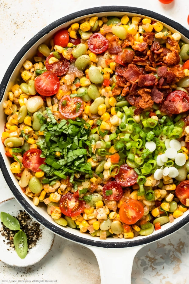 Overhead photo of corn and lima bean side dish topped with bacon, basil and scallions in a white skillet.