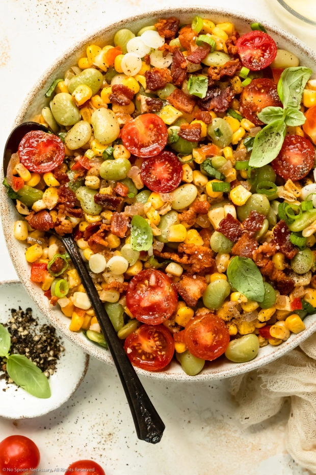 Overhead photo of succotash with bacon in a white serving bowl with a spoon inserted into the dish.