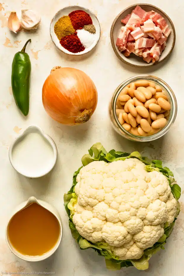 Overhead photo of a head of cauliflower, broth, white beans, a yellow onion, jalapeno, and seasonings neatly arranged on a kitchen counter.