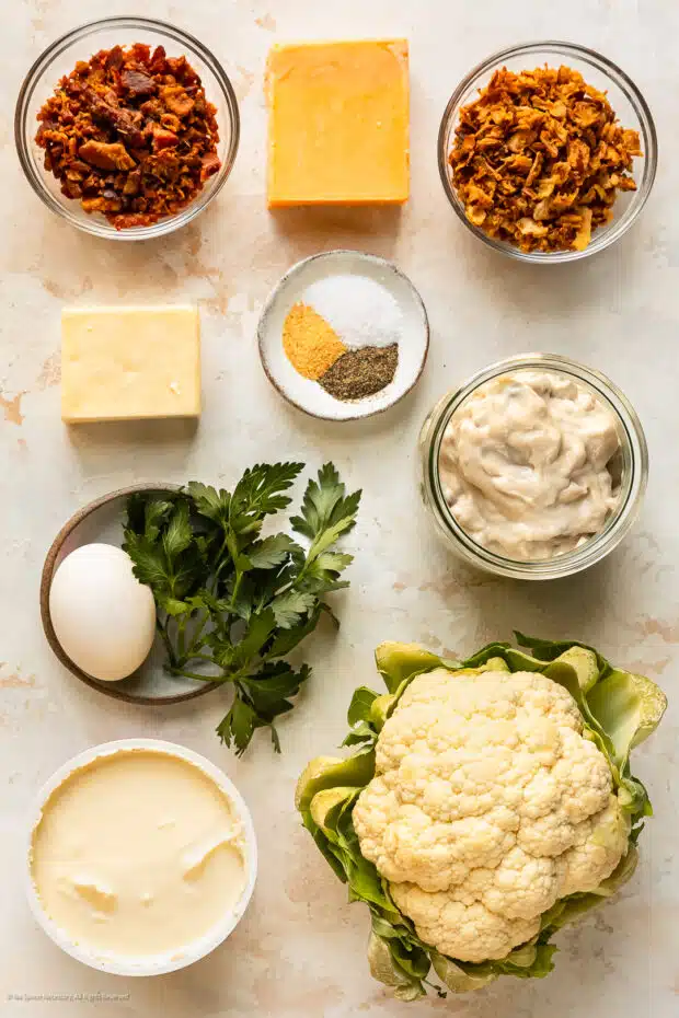 Overhead photo of a head of cauliflower, blocks of cheese, crumbled bacon, condensed soup, ground seasonings, and fresh herbs neatly organized on a kitchen counter.