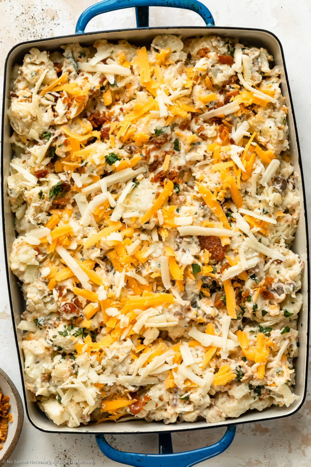 Overhead photo of an unbaked cauliflower casserole topped with shredded cheese in a blue baking dish.