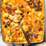 Overhead photo of cheesy cauliflower casserole loaded with bacon and crispy onions in a large blue baking dish with a serving spoon inserted into the casserole.