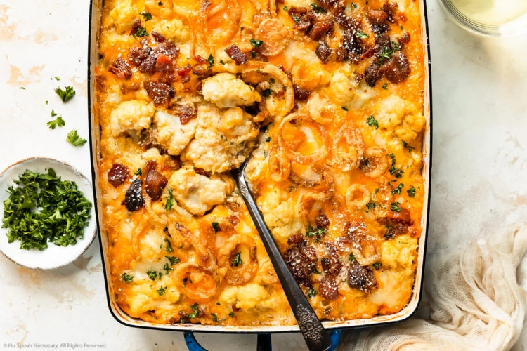 Overhead photo of cheesy cauliflower casserole loaded with bacon and crispy onions in a large blue baking dish with a serving spoon inserted into the casserole.