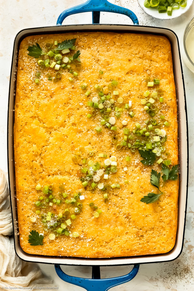 Overhead photo of creamed corn casserole topped with grated parmesan and sliced scallions in a blue baking dish.