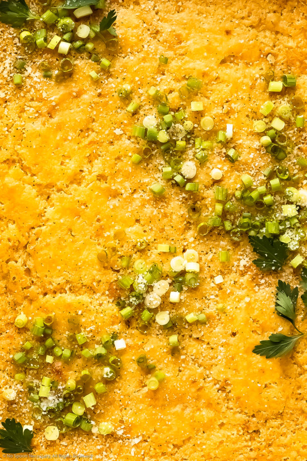 Overhead, close-up photo of the top of a homemade corn casserole garnished with grated parmesan and sliced scallions.