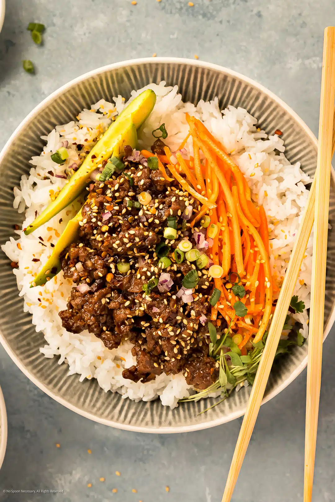 Overhead photo of white rice topped with ground beef stir fry, sliced avocados, and julienne carrots in a serving bowl.