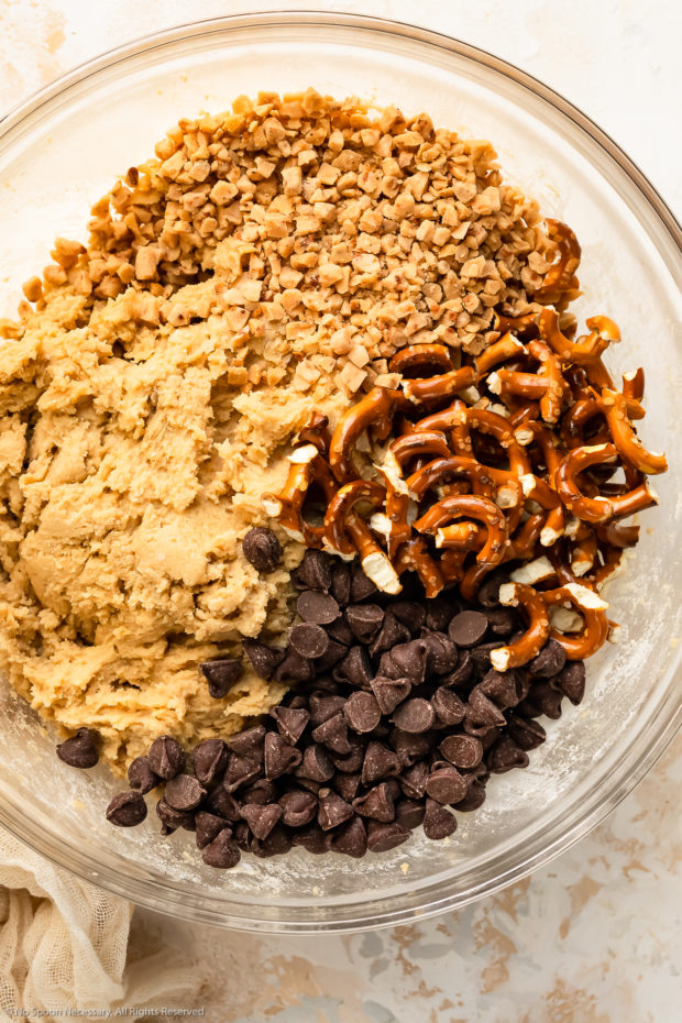 Overhead photo of raw cookie dough topped with toffee bits, crushed pretzels and chocolate chips in a glass bowl.