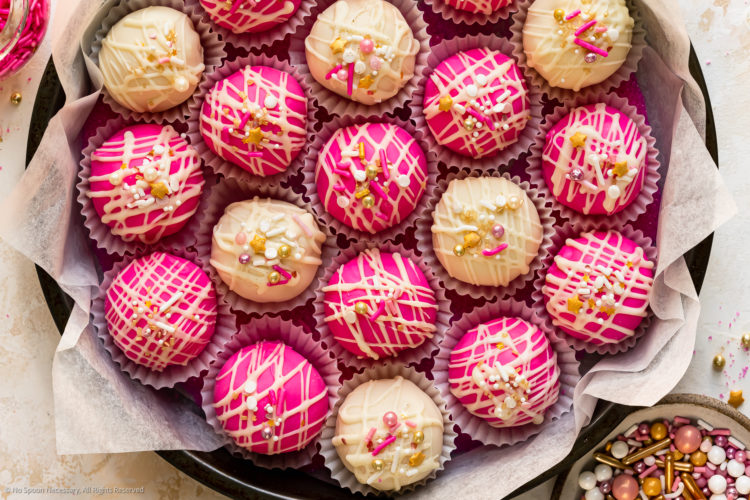 Overhead photo of hot pink and white homemade cake bites in a cookie tin with a bowl of sprinkles next to the tin.
