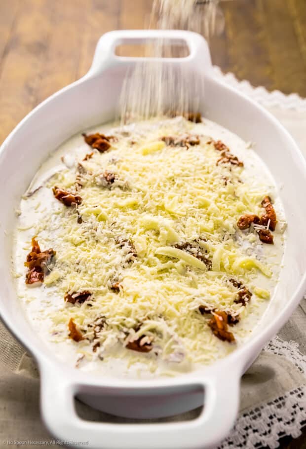 Action photo of grated cheese being sprinkled over chicken with sundried tomatoes and cream sauce in a casserole dish.