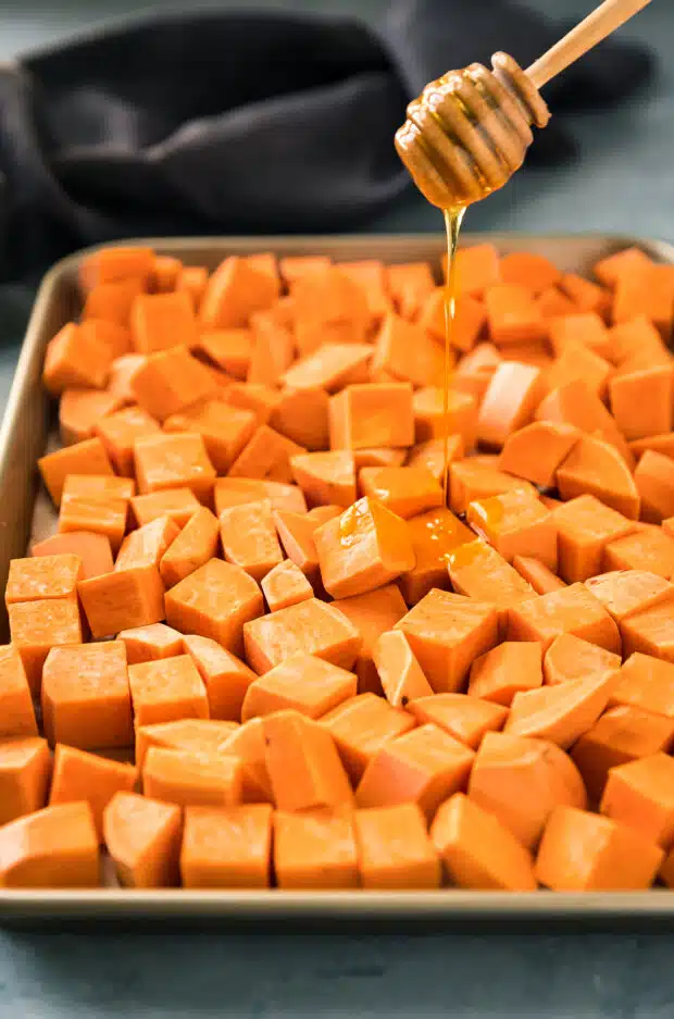 Action photo of honey being drizzled over a cubes of sweet potato cubes for roasting and using in soup.