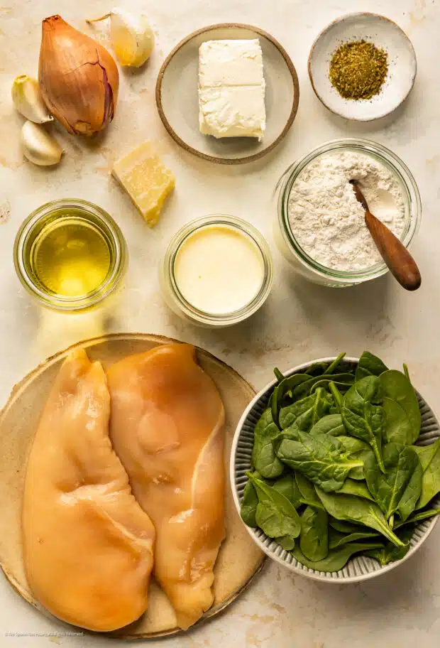 Overhead photo of all the ingredients for florentine chicken (chicken breasts, baby spinach, cream cheese, wine, aromatics and seasonings) neatly arranged on a kitchen counter.