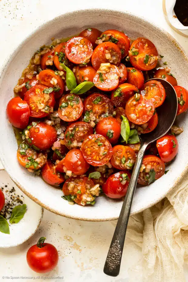Overhead photo of marinated cherry tomatoes with fresh basil in a white serving bowl with a spoon.