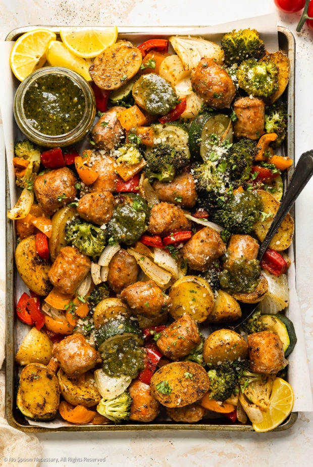 Overhead photo of cooked sausages, peppers and potatoes on a sheet pan with a jar of pesto.
