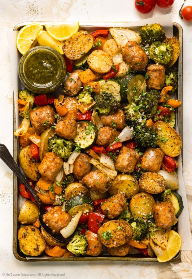 Overhead photo of cooked sausage and veggies on a sheet pan with a jar of pesto sauce.