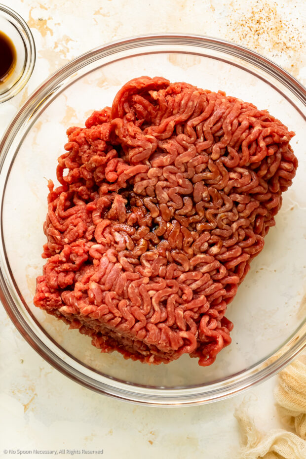 Overhead photo of raw ground beef drizzled with Worcestershire and soy sauce in a glass bowl.