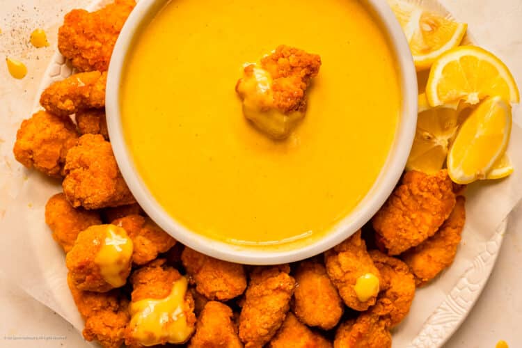 Overhead photo of a chicken nugget resting in a bowl of honey mustard dipping sauce with more nuggets surrounding the bowl.