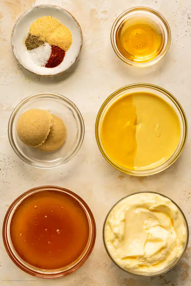 Overhead photo of honey, mustard, brown sugar, mayonnaise, and seasonings organized into individual bowls on a kitchen counter.