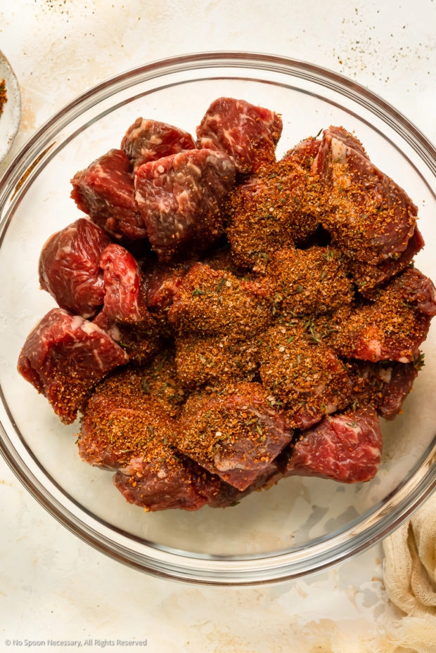 Overhead photo of raw steak bites sprinkled with seasonings in a large mixing bowl.