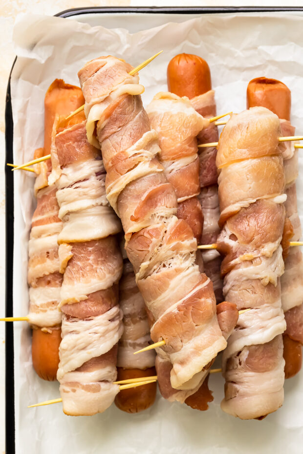 Overhead photo of a stack of raw hot dogs wrapped with strips of bacon on a parchment-lined tray.