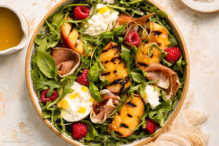 Overhead photo of grilled peach salad with burrata, prosciutto, and arugula in a large serving bowl.