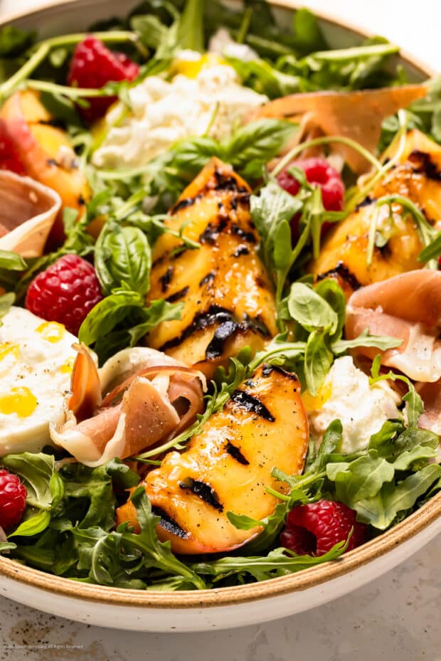 Angled photo of a burrata and peach salad with arugula in a white bowl.