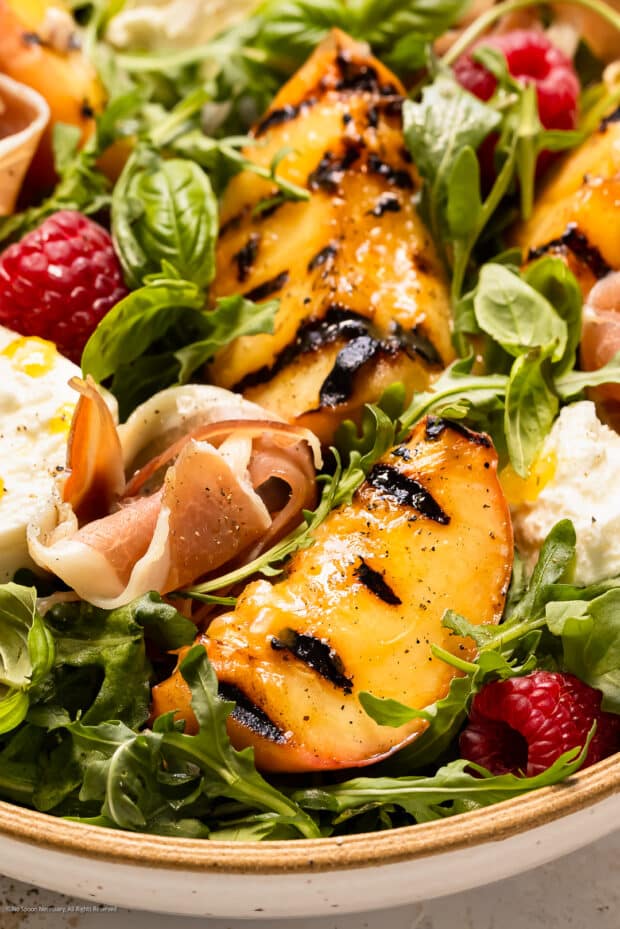 Close-up photo of a grilled peach wedge on a bed of arugula lettuce with torn burrata.