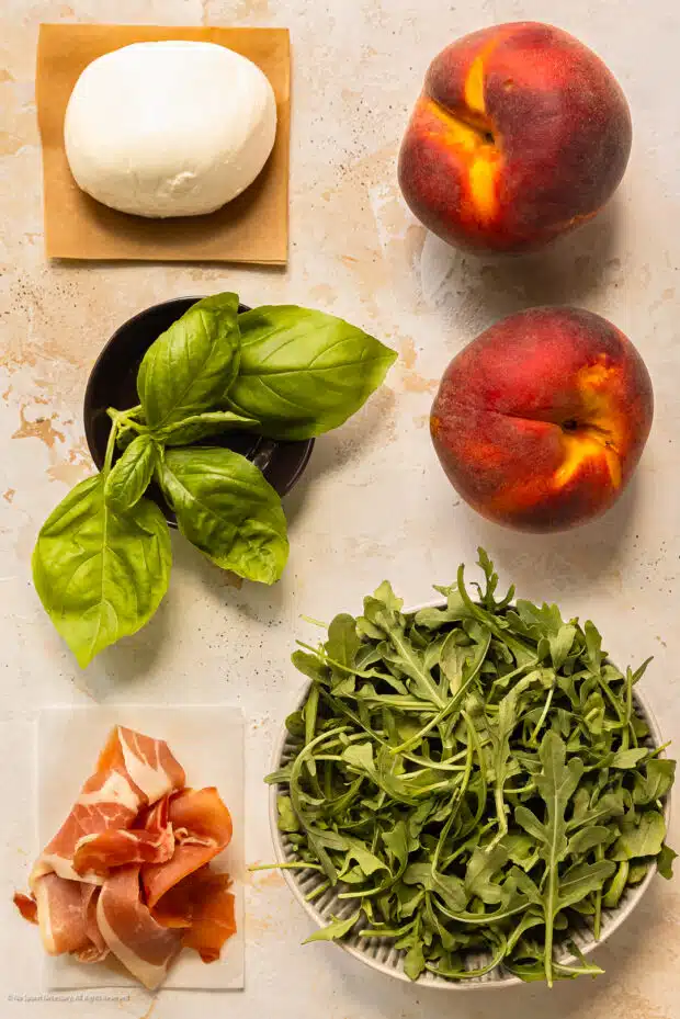 Overhead photo of a ball of burrata, two peaches, basil, arugula, and prosciutto slices neatly arranged on a kitchen counter.