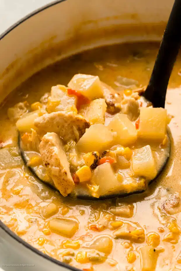Angled action photo of a person ladling chicken chowder from a white pot.