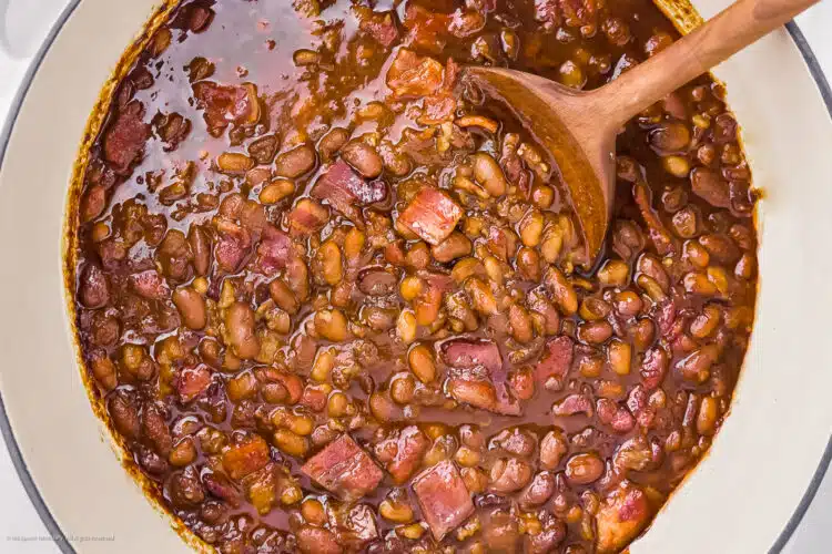 Overhead photo of baked beans with bacon in a large white pot with a wood serving spoon.