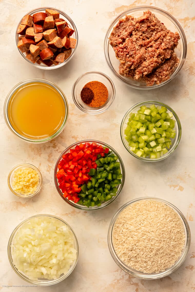 Overhead photo of ground sausage, andouille sausage, rice grains, chicken broth, and seasoning neatly arranged on a kitchen counter.